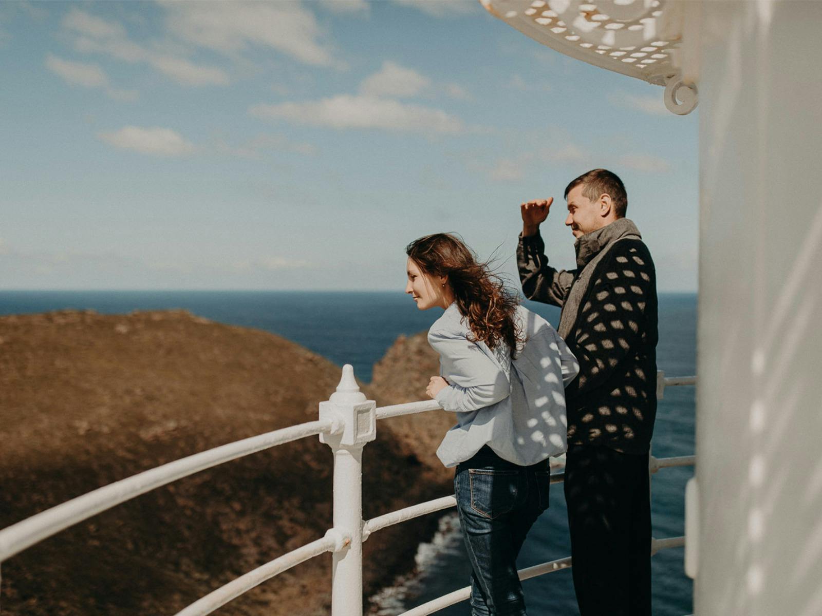Couple on top of lighthouse looking at mountains and ocean beyond.