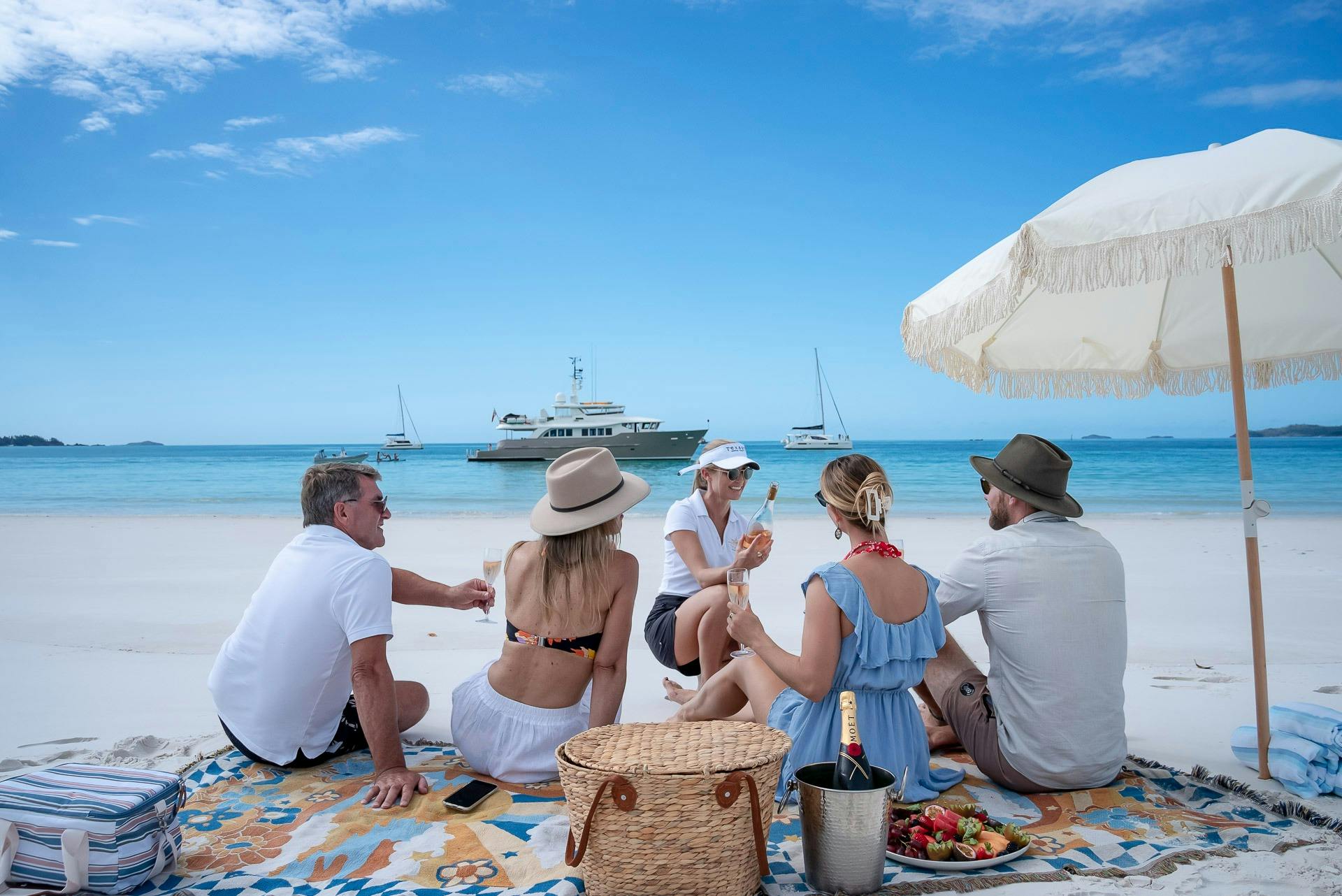 Two couples sitting on picnic rug with umbrella being served champagne on Whitehaven Beach
