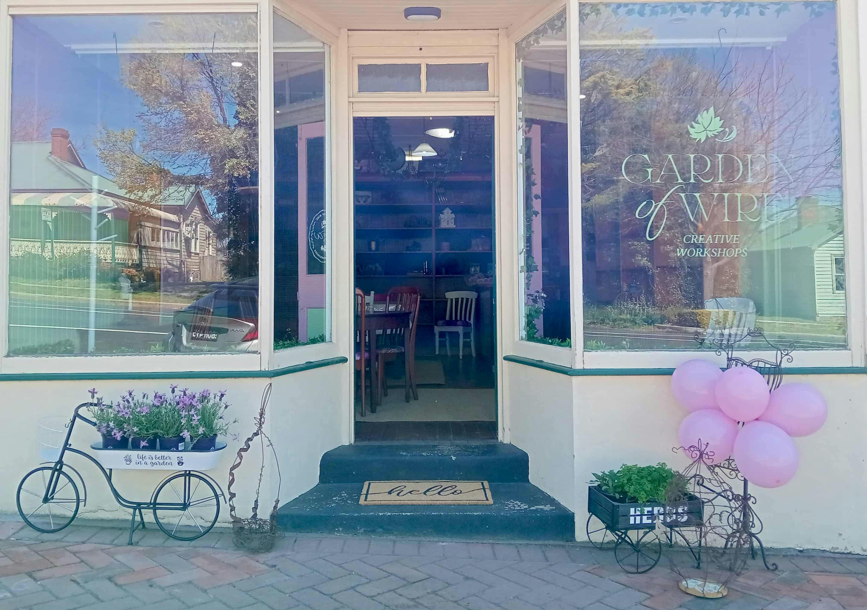 Street view of shop with 2 bay windows and recessed door. Door is open to welcome guests.