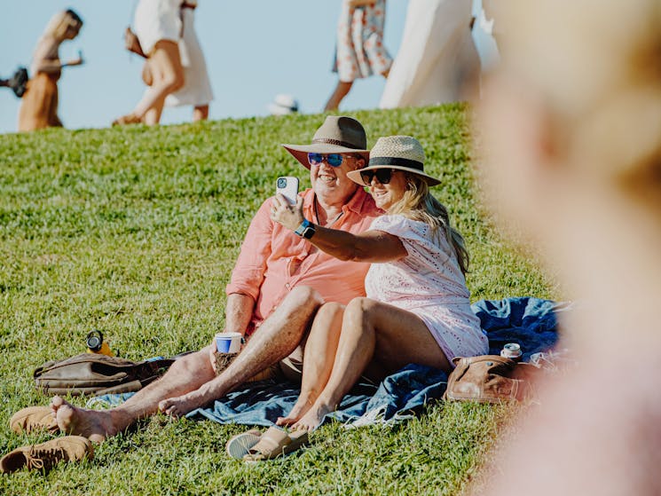 two patrons, a male and female, sit on a picnic blanket smiling  while taking a photo of themselves