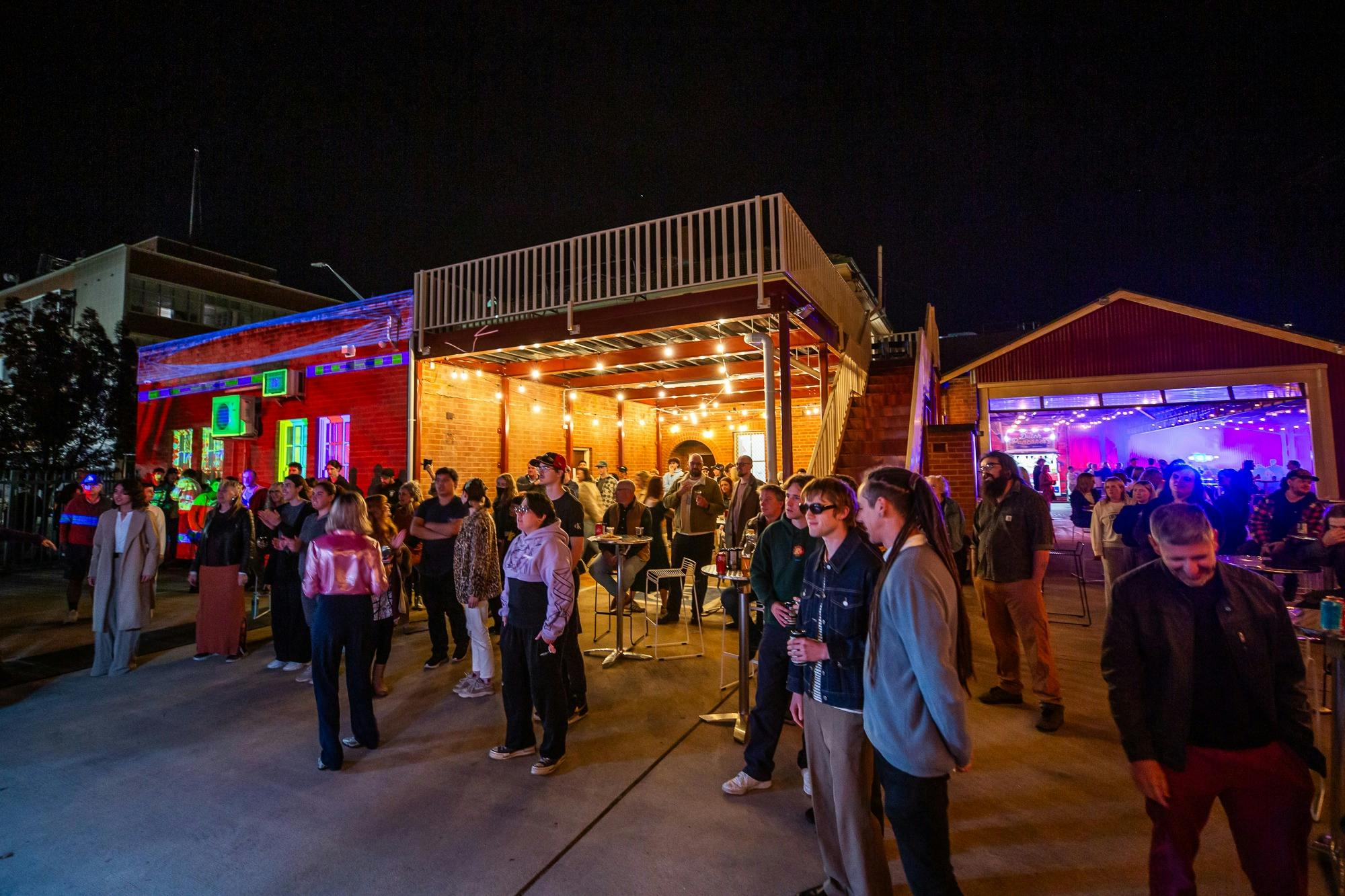 An open, outdoor event area with colourful light installation and guests watching live music.