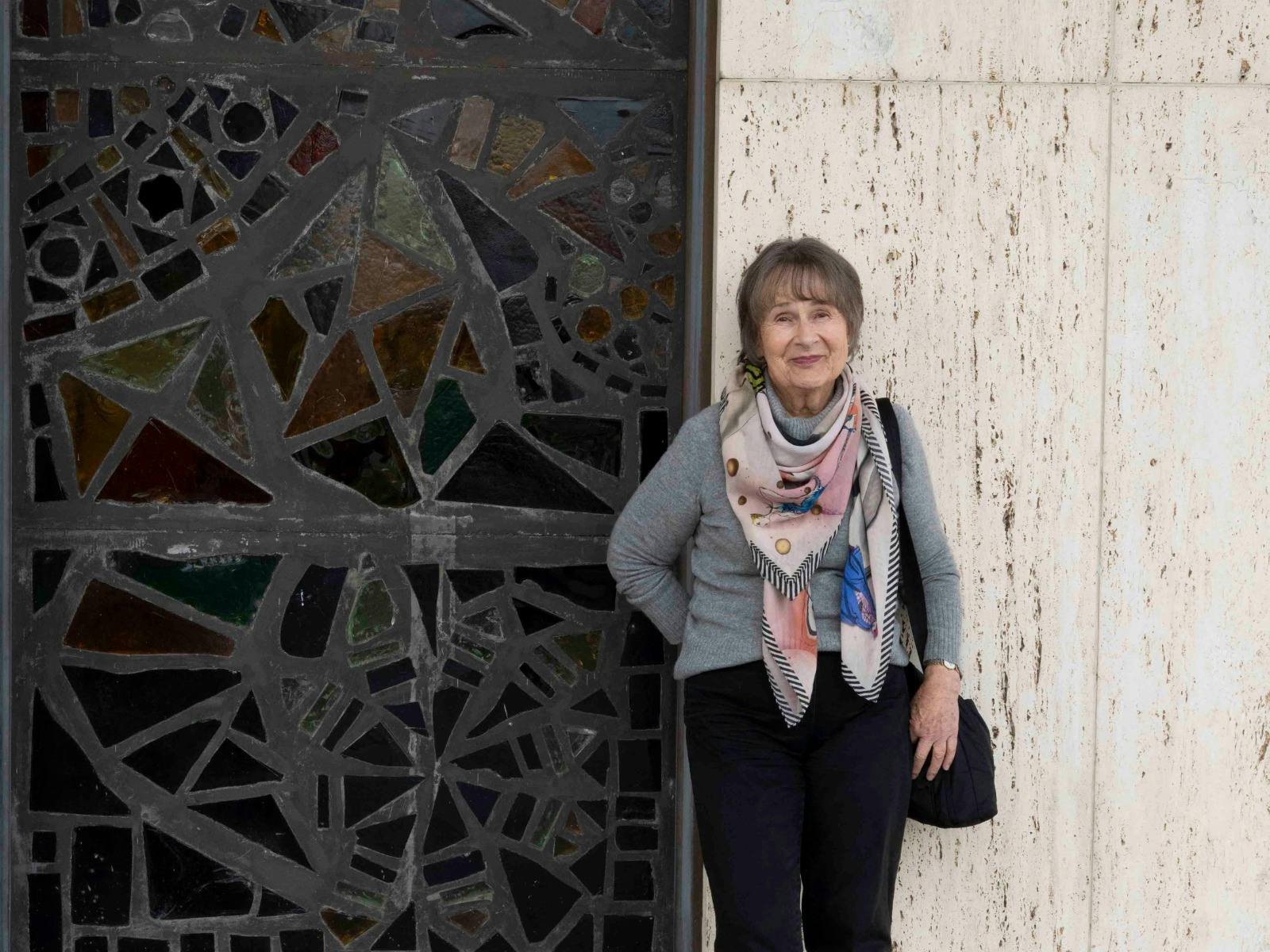 An woman with short gray hair stands smiling beside a decorative mosaic wall.
