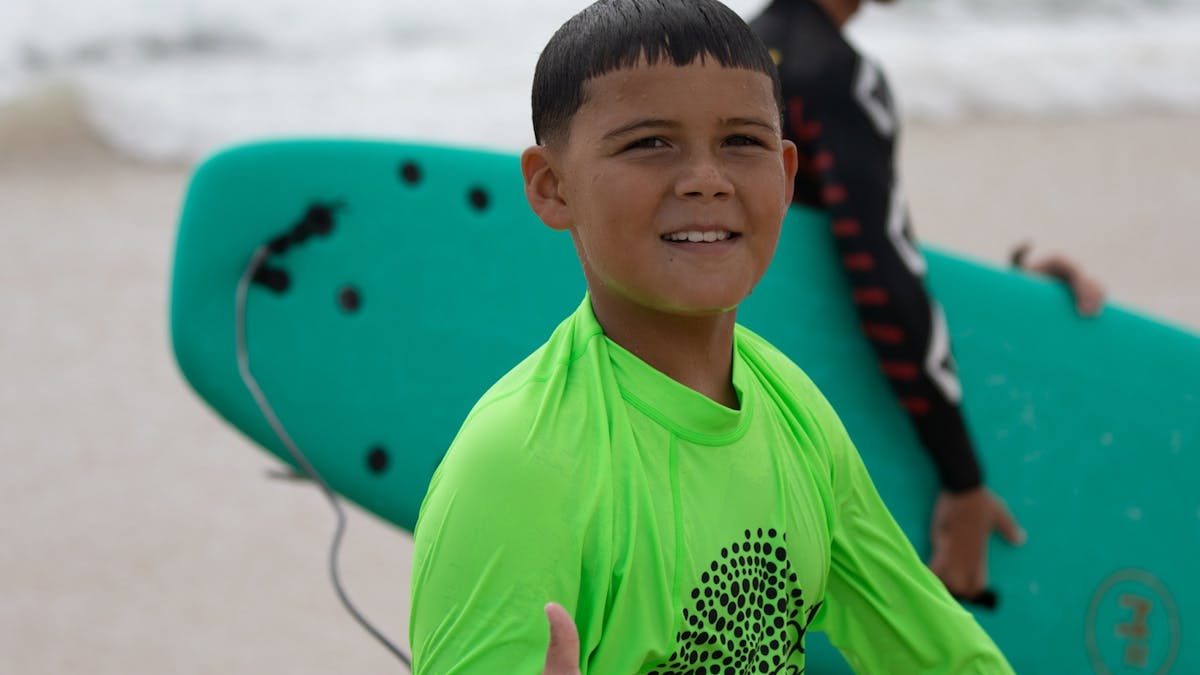 Boy gives thumbs up after surf lesson, green surfboard in background