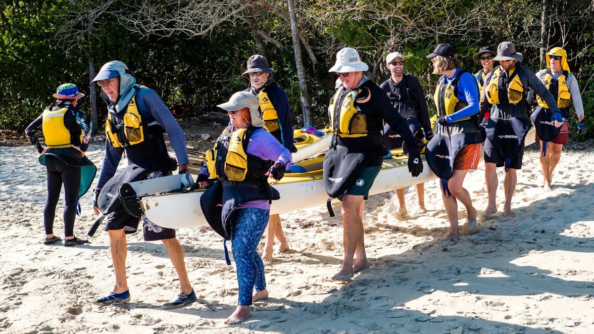 10 people work together to carry a sea kayak down a beach to the waters edge. They are smiling.