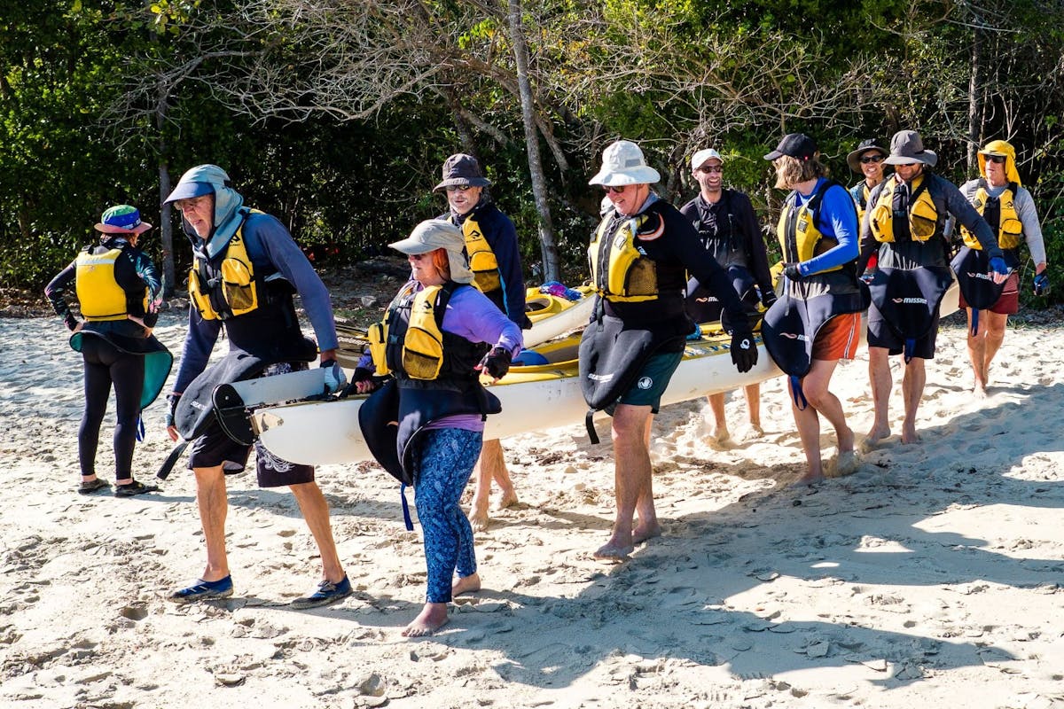 10 people work together to carry a sea kayak down a beach to the waters edge. They are smiling.