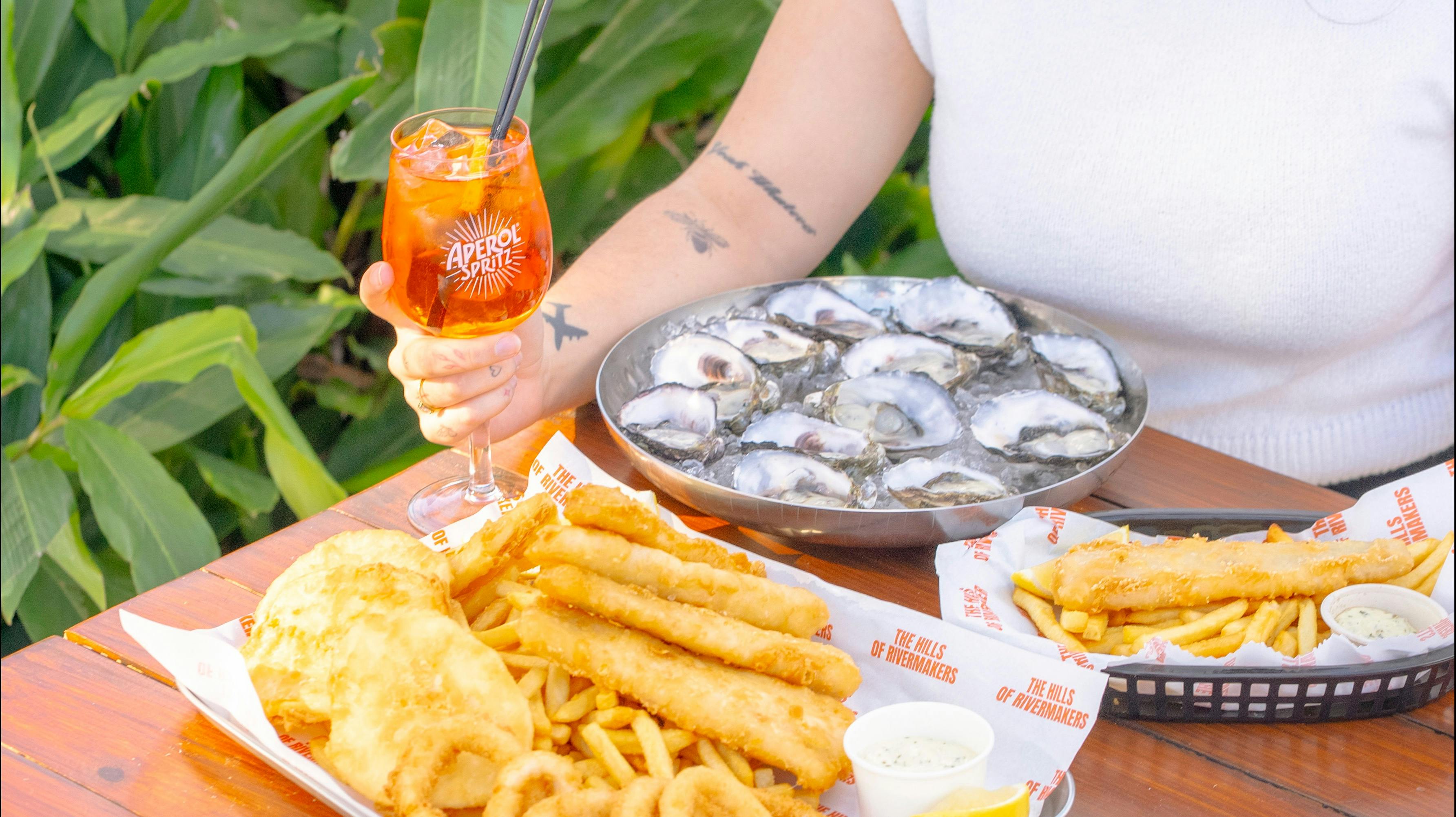 Person enjoying oysters, fish and chips, and an Aperol Spritz at an outdoor table.