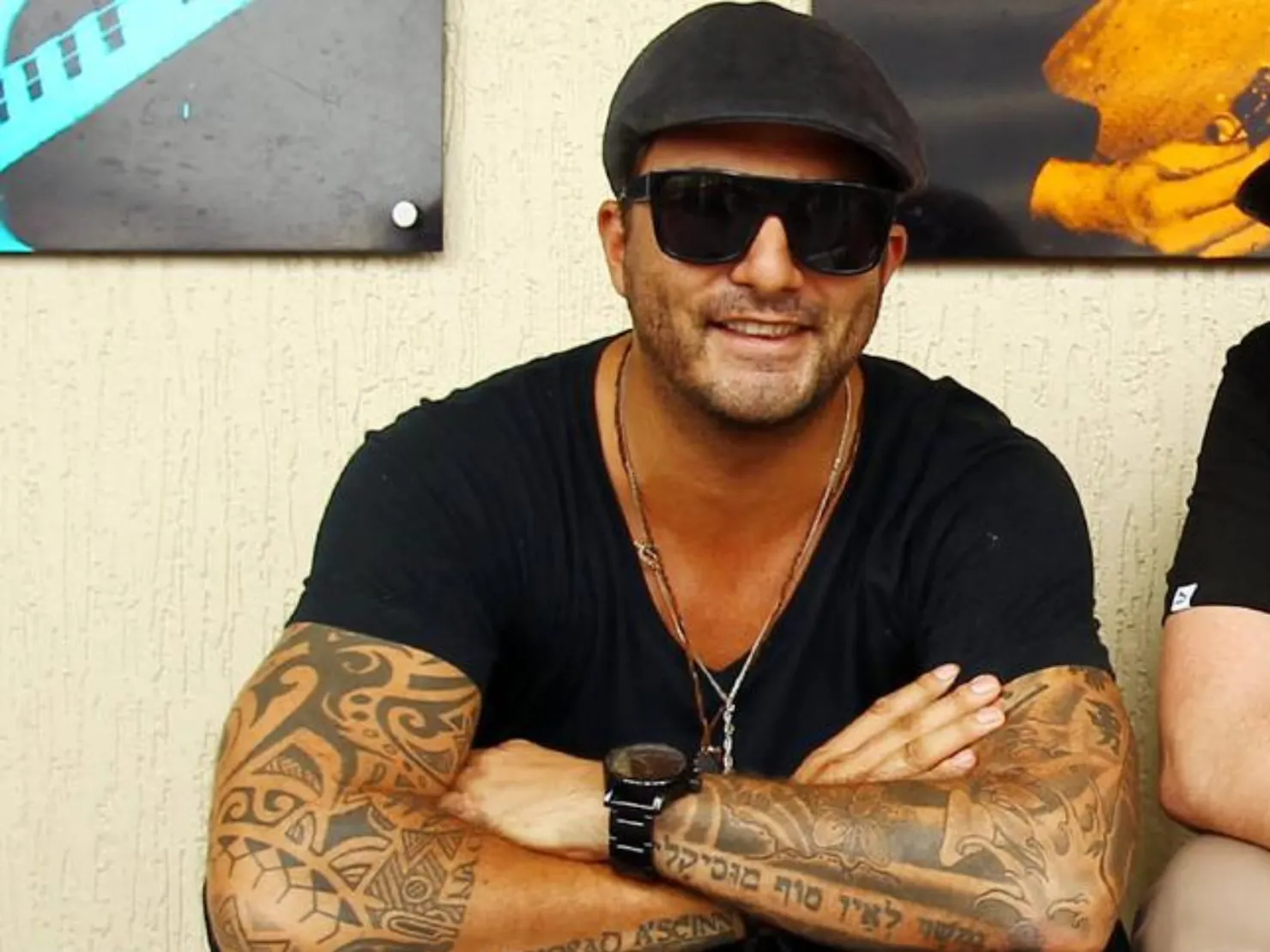 Man sitting with his arms crossed over he's legs smiling with black glasses and hat on
