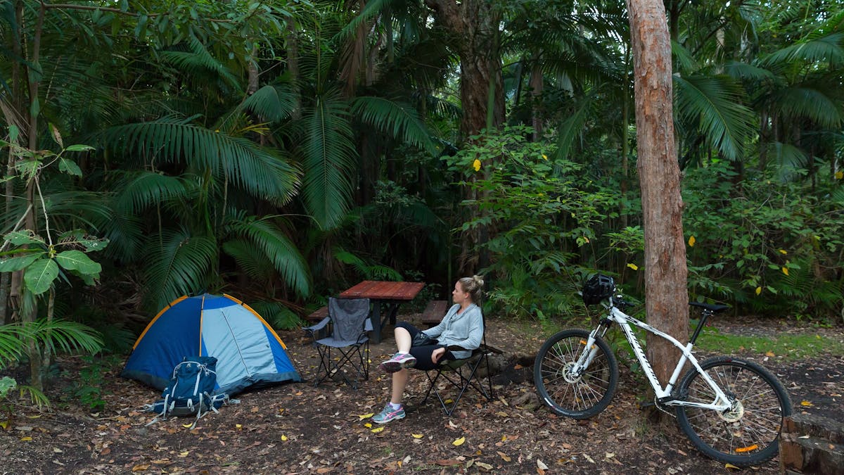 Person sits near her small tent and mountain bike surrounded by tall forest.