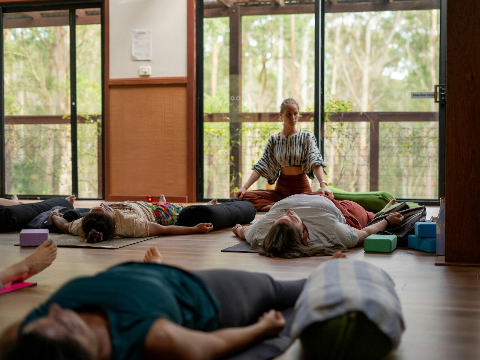 A yoga teached sitting cross legged in a class of people lying on the ground in meditation