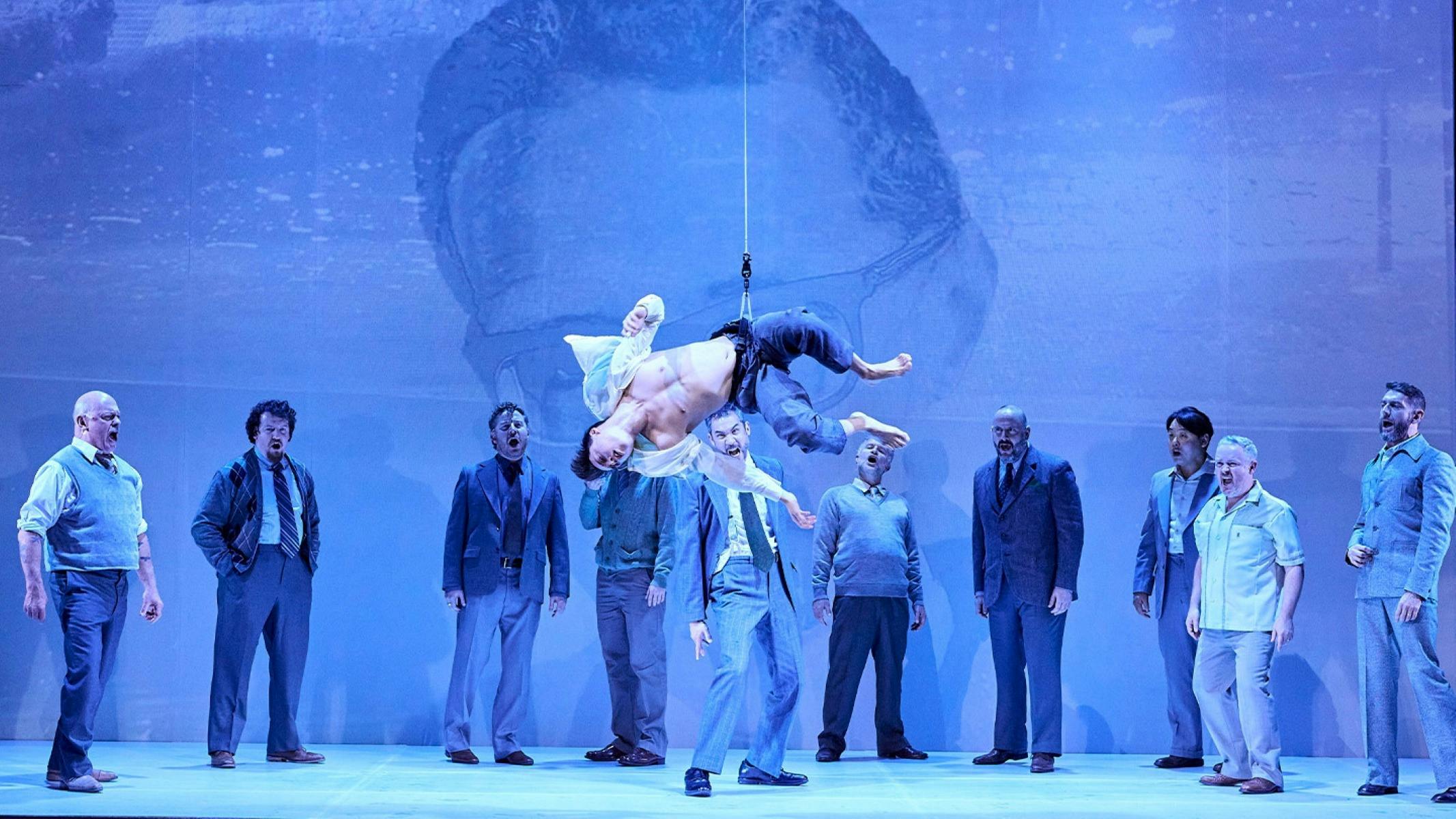 Performer suspended above a stage as an ensemble surrounds them in a dramatic opera scene.