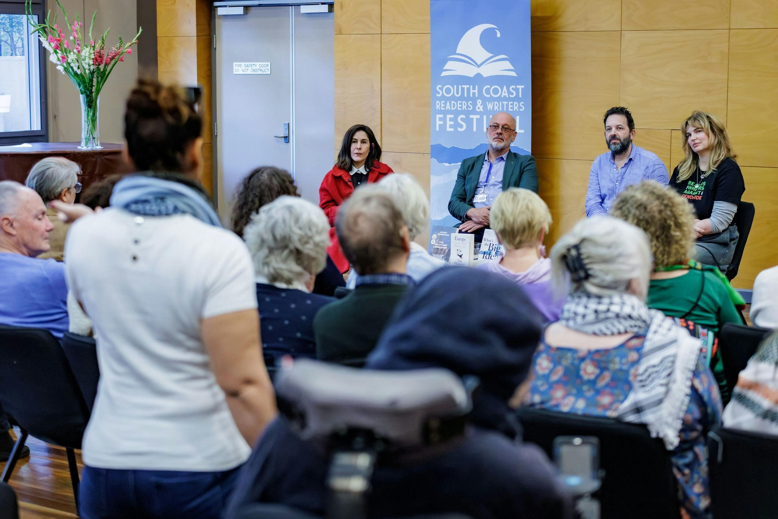 Four people sitting on chairs in front of a crowd, listening to a question from the audience