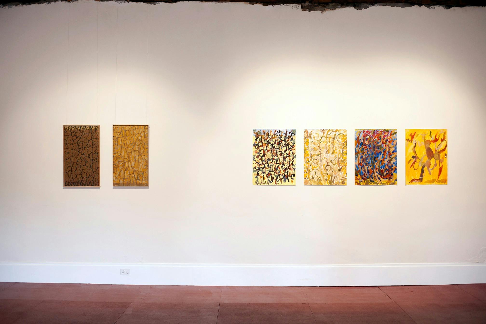 Colourful paintings and drawings are hung on a white wall with a red floor in the bottom of frame.