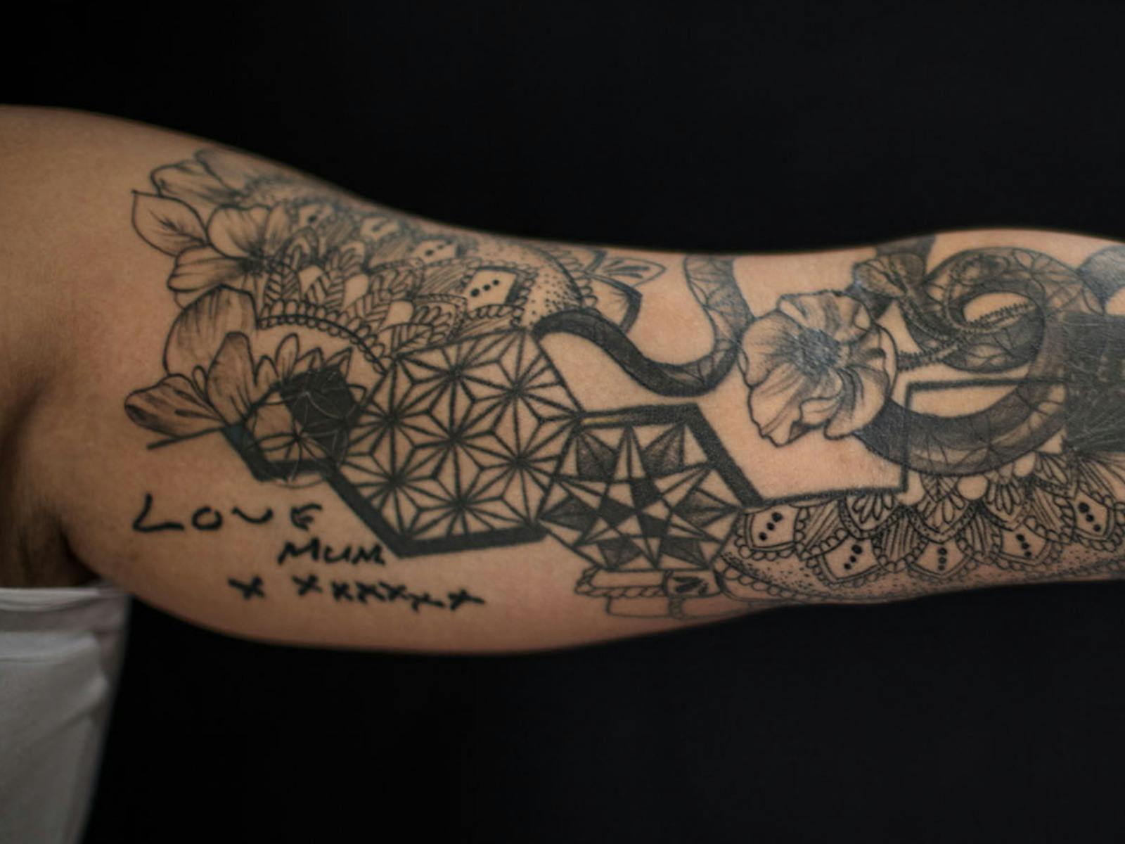 Black and grey tattoo on an arm featuring geometric patterns, flowers, and handwriting.