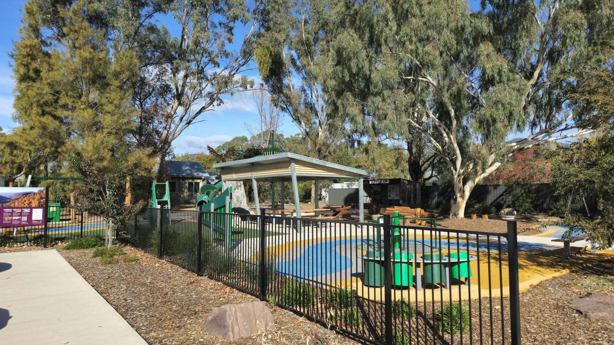 Black fence running along the front of a playground. Trees and blue sky in background