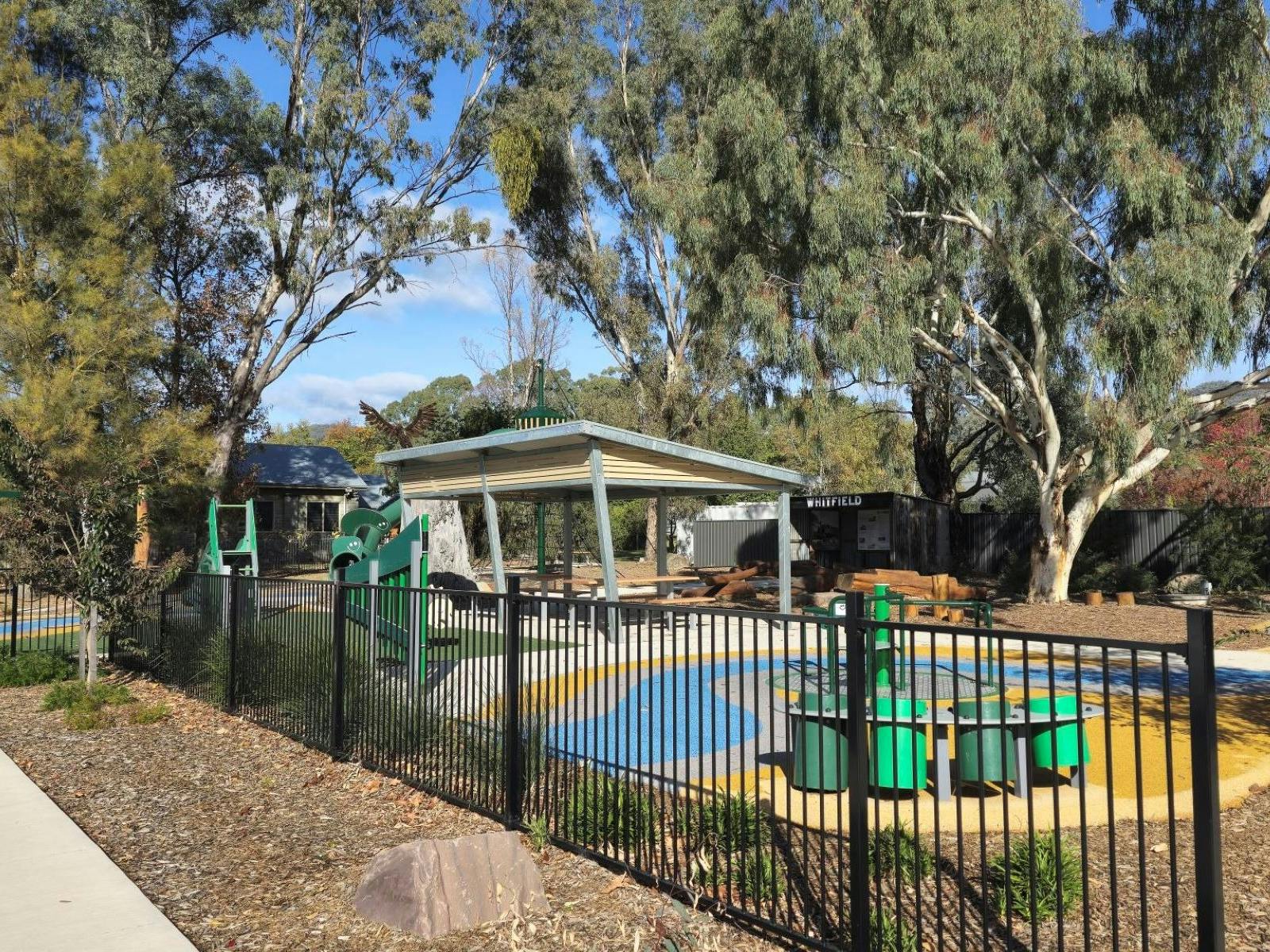 Black fence running along the front of a playground. Trees and blue sky in background