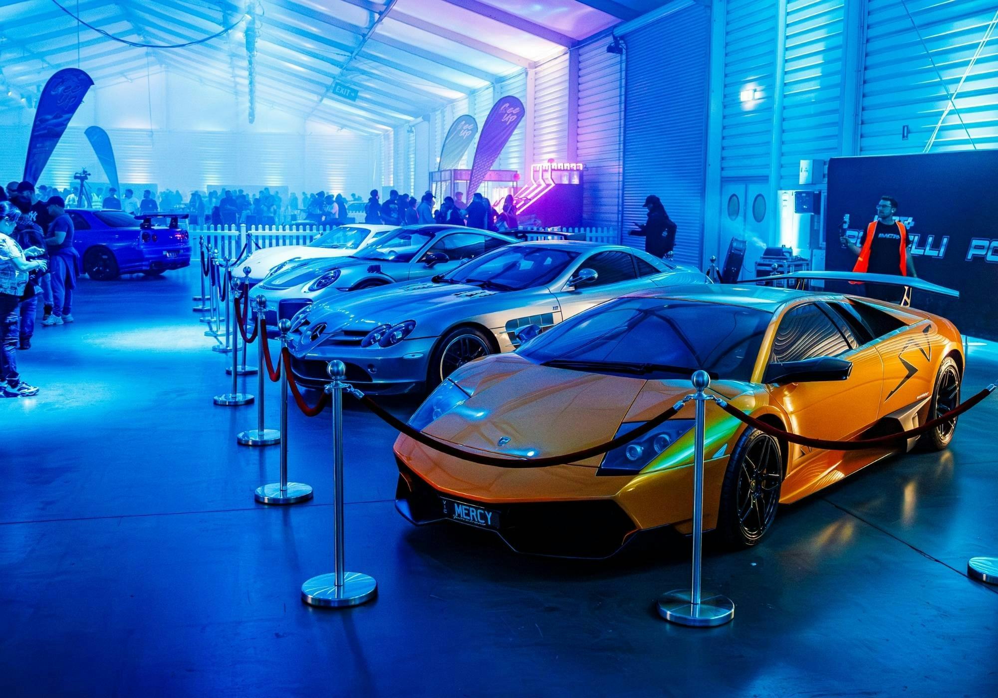Exotic supercars displayed behind ropes under blue lighting inside Portelli World.