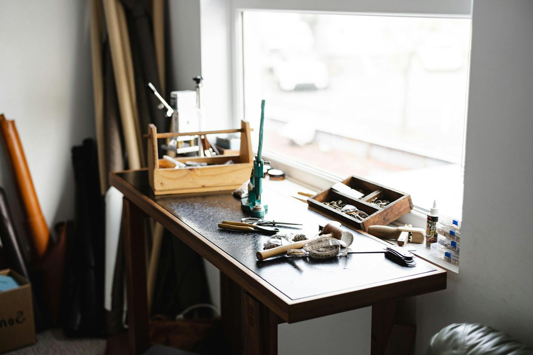 Leather crafting workbench by a window, with tools, hardware, and materials scattered across surface