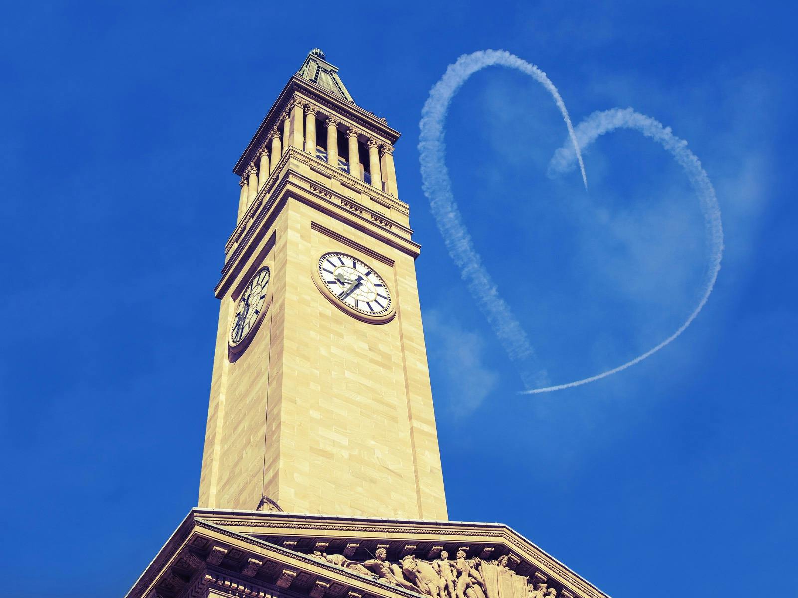 A sandstone clock tower against a bright blue sky with a heart beside it in sky-writing.
