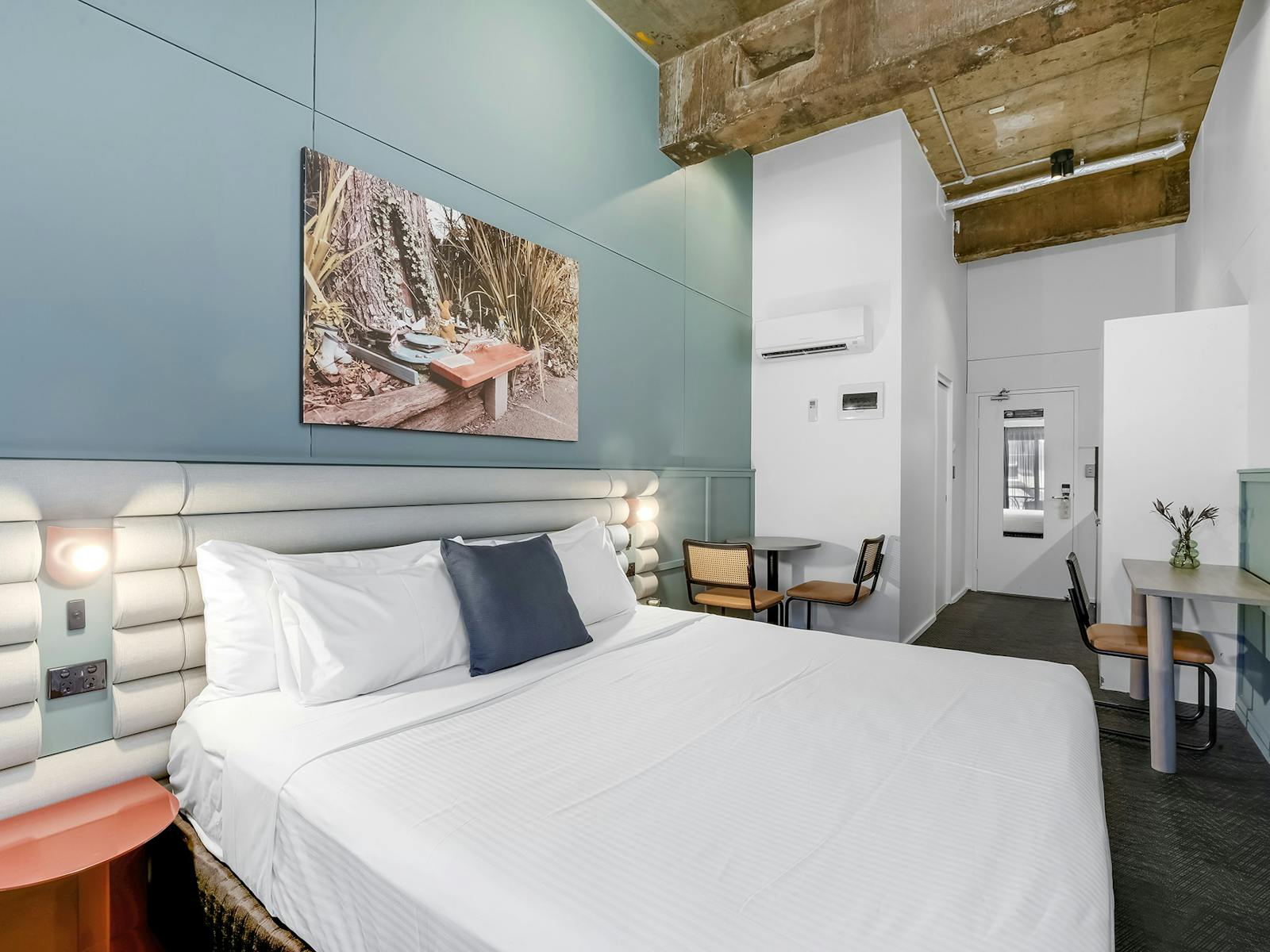 높은 산업용 천장과 책상이 있는 스튜디오에 깨끗한 흰색 시트가 깔린 갓 정돈된 호텔 침대가 있습니다.