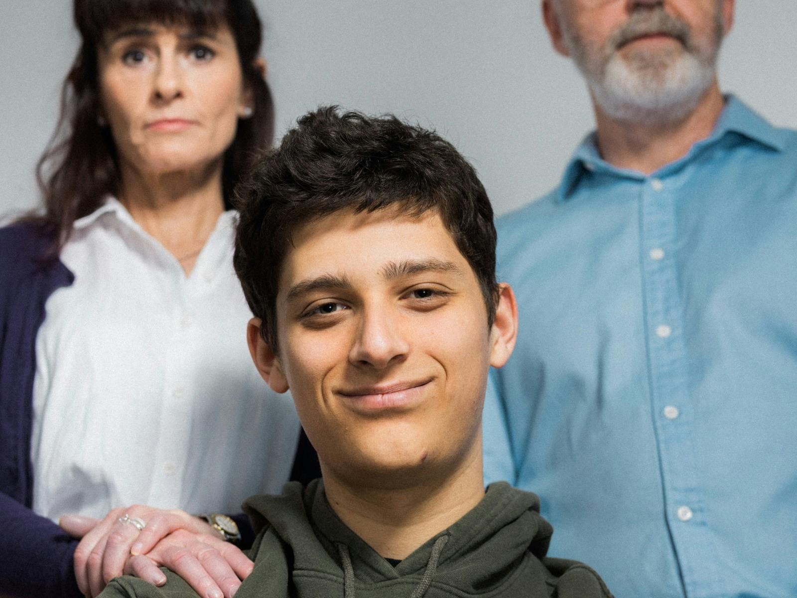 A young man smirking at the camera in the foreground, with two adults in the background, concerned