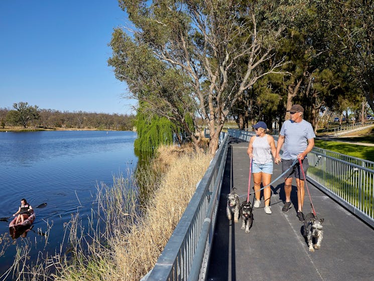 Two people walking dogs on boardwalk on right with kayaker on Lake Inverell on left