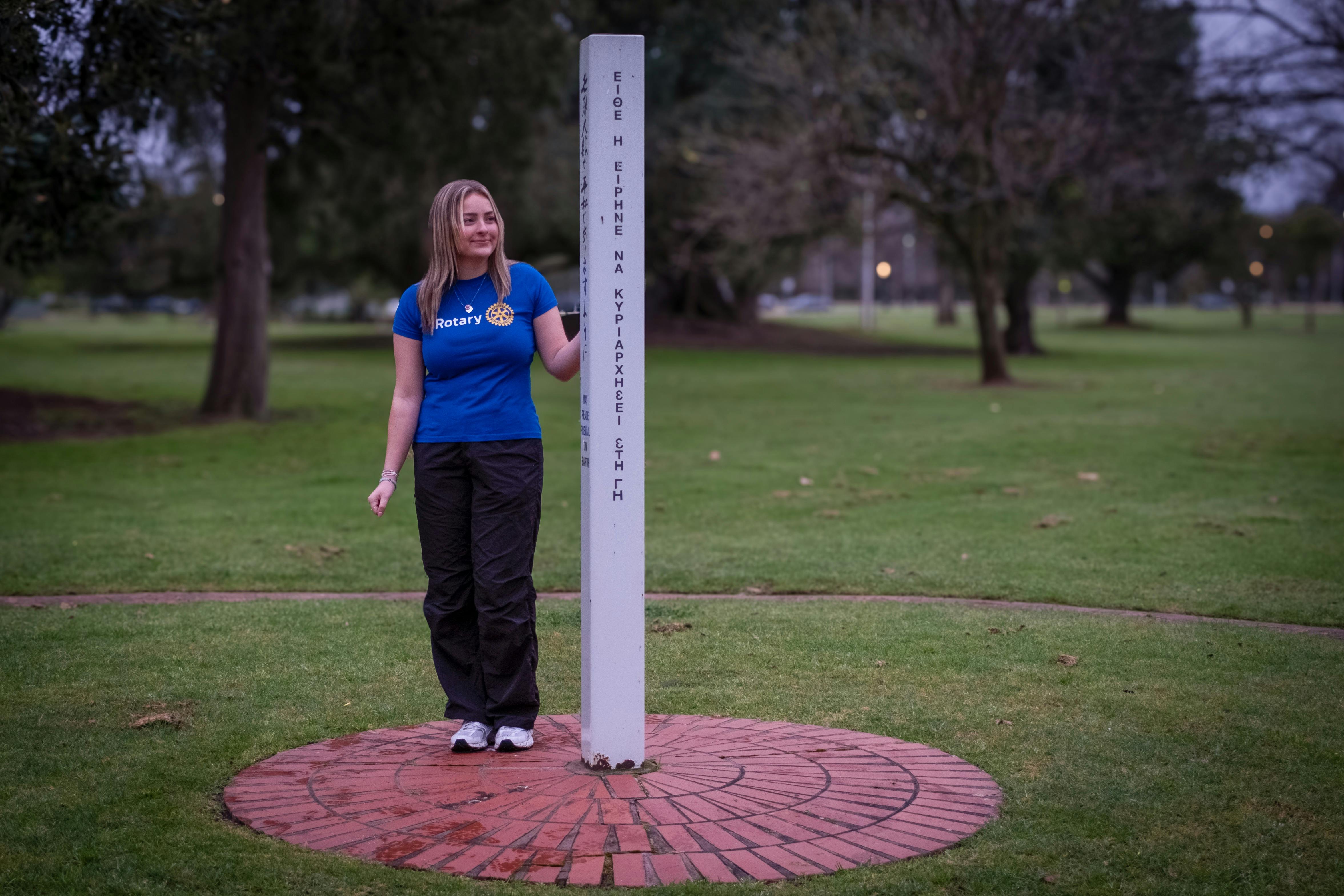 a white pole with the words May Peace Prevail on Earth with a woman standing next to it in a park