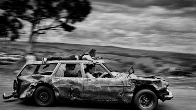 black & white image of a banged-up car speeding past with passengers and one man on the car roof