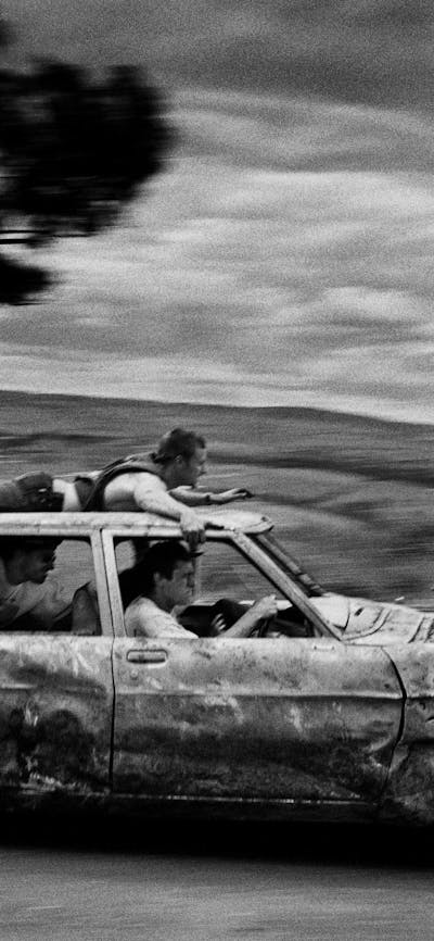 black & white image of a banged-up car speeding past with passengers and one man on the car roof