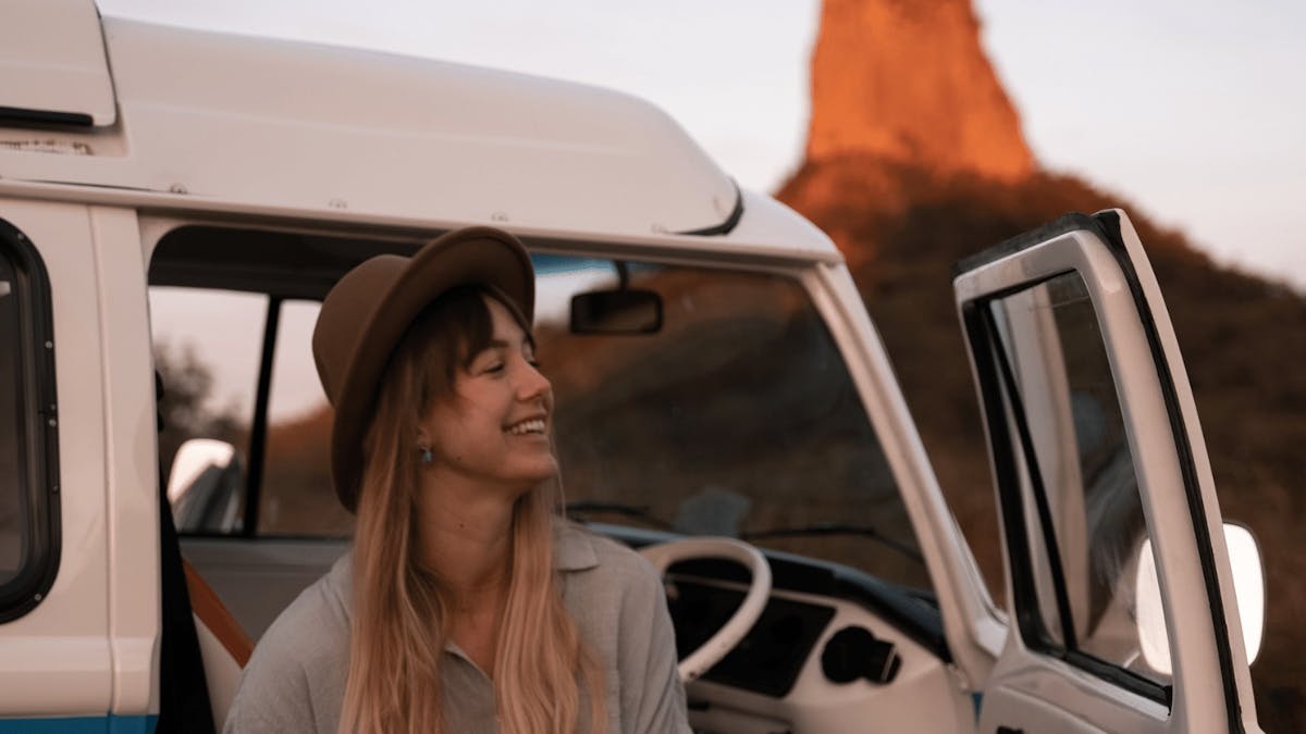 Young female standing outside the door of a blue kombi in front of Glass House Mountain