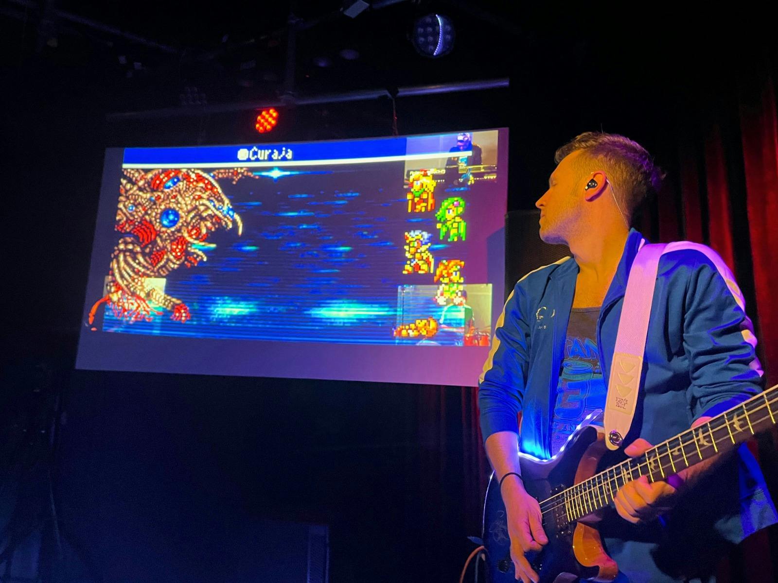 A man playing a guitar standing next to a TV screen with a video game