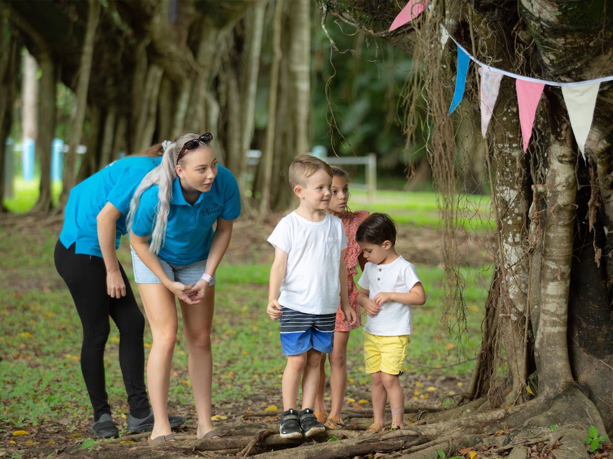 Image of a Nanny playing hide and seek with two children by a tree with bunting in it