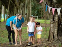 Image of a Nanny playing hide and seek with two children by a tree with bunting in it