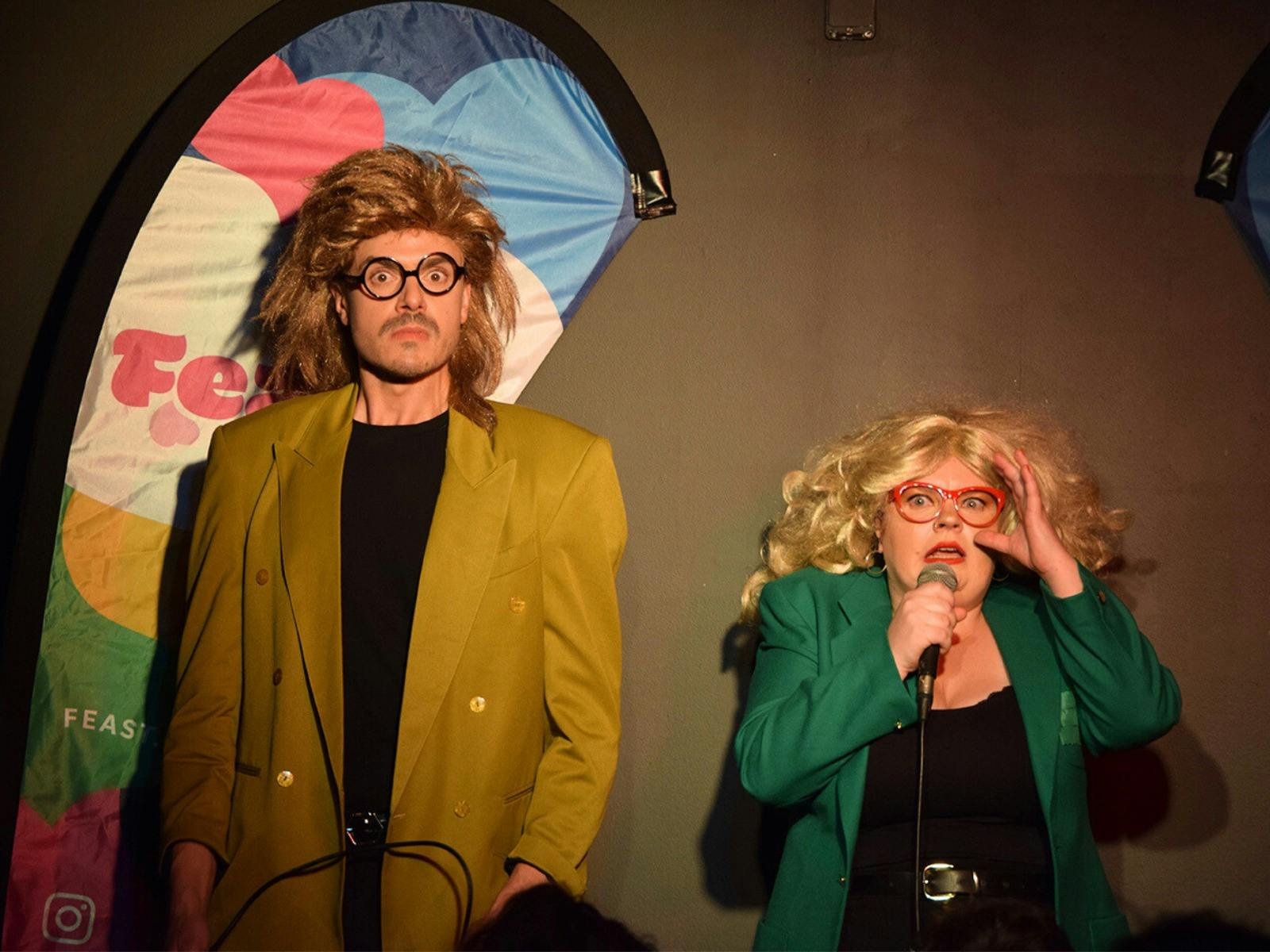 Two performers wearing glasses and blonde wigs, making funny serious faces.