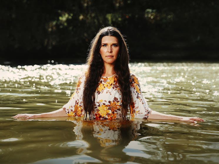 Woman standing waist deep in water, arms resting on surface, wearing floral dress in autumnal tones.