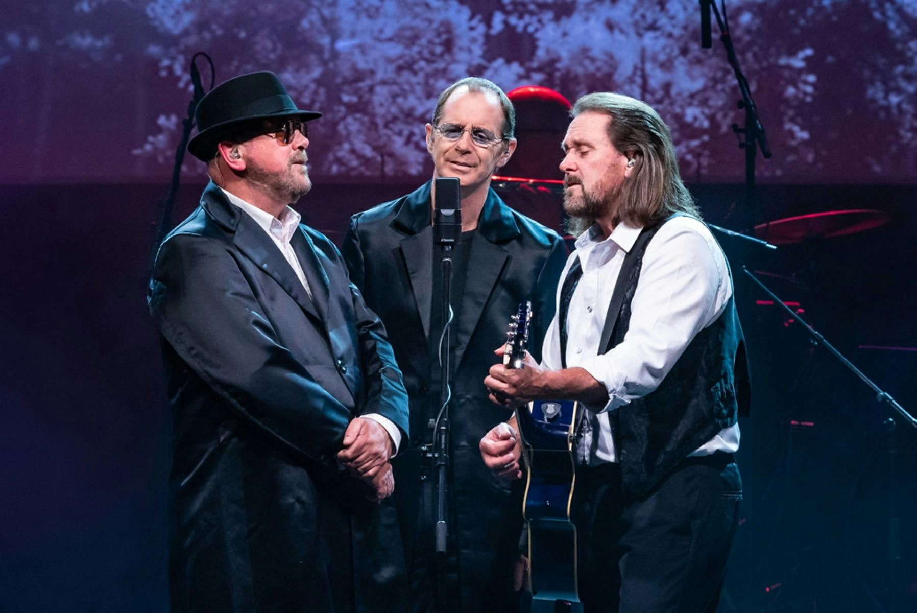 Three men singing into one microphone on stage