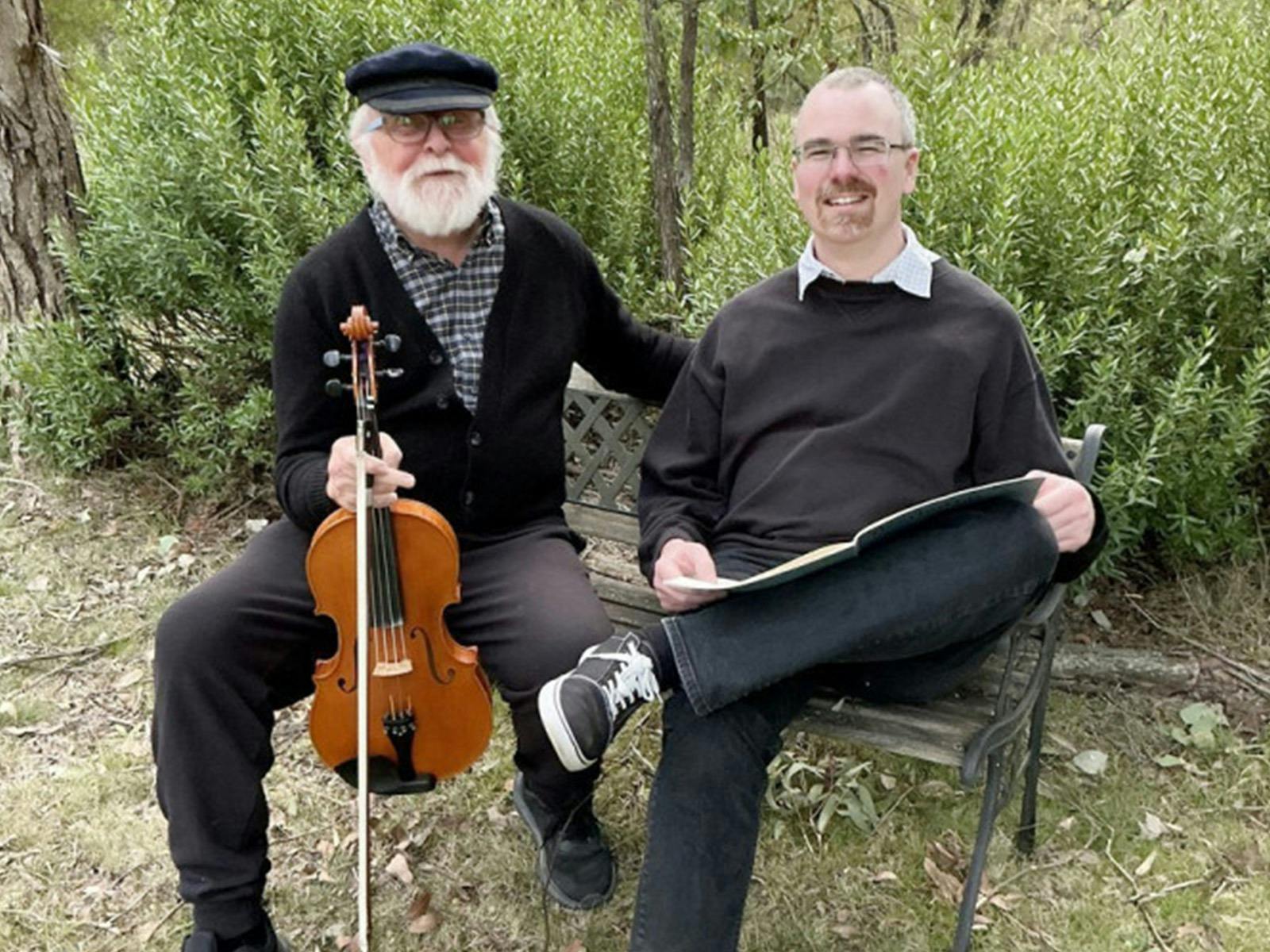 Two elderly men sitting on a bench in nature, one holding a viola