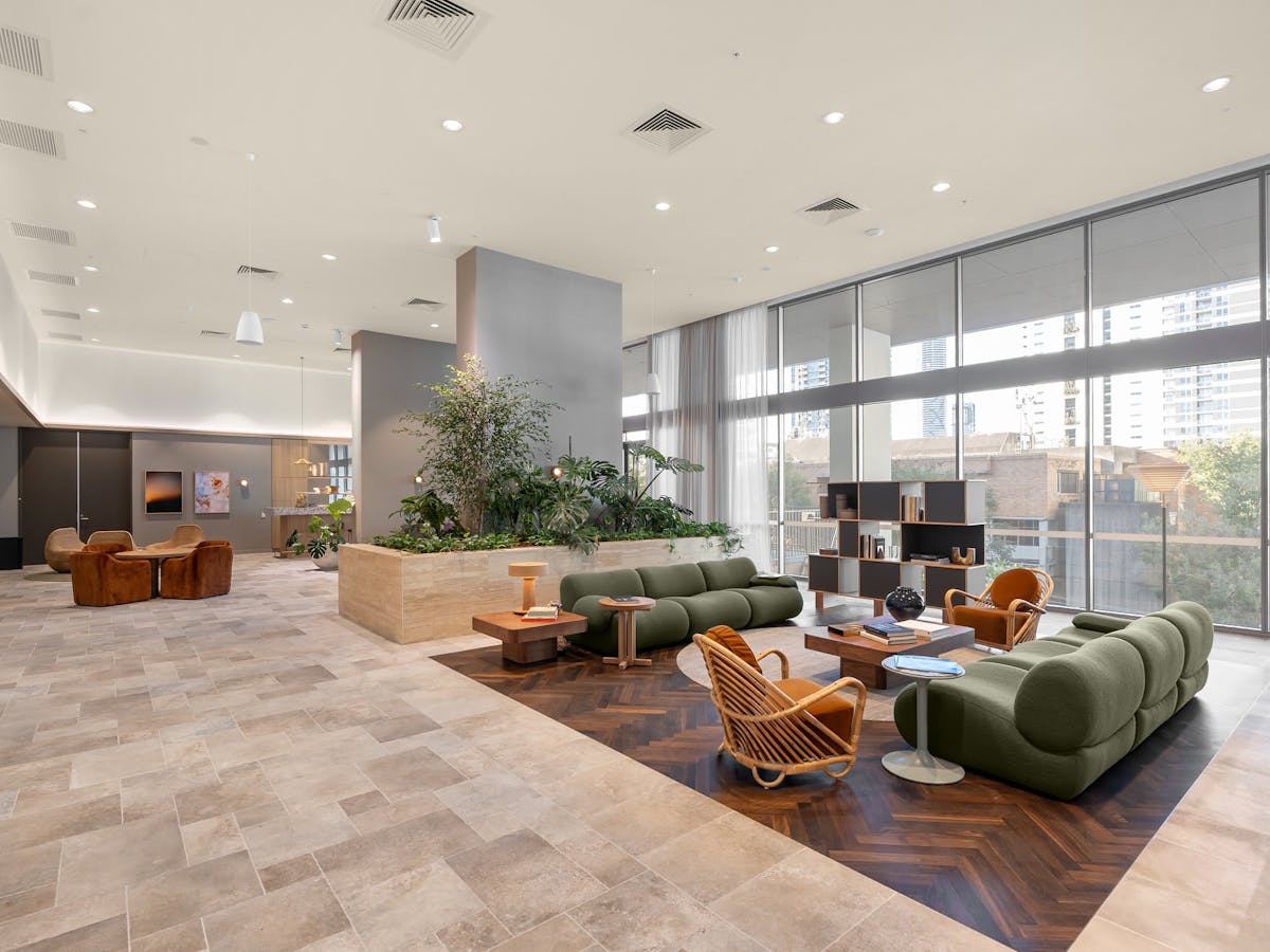 Spacious communal lounge area with green couches, plants, floor to ceiling windos and orange chairs.