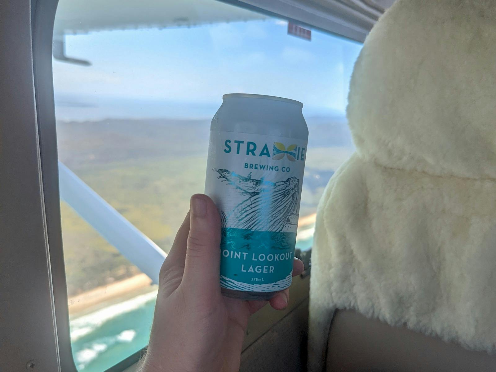 A Straddie Brewing Co Point Lookout Lager against an aircraft window