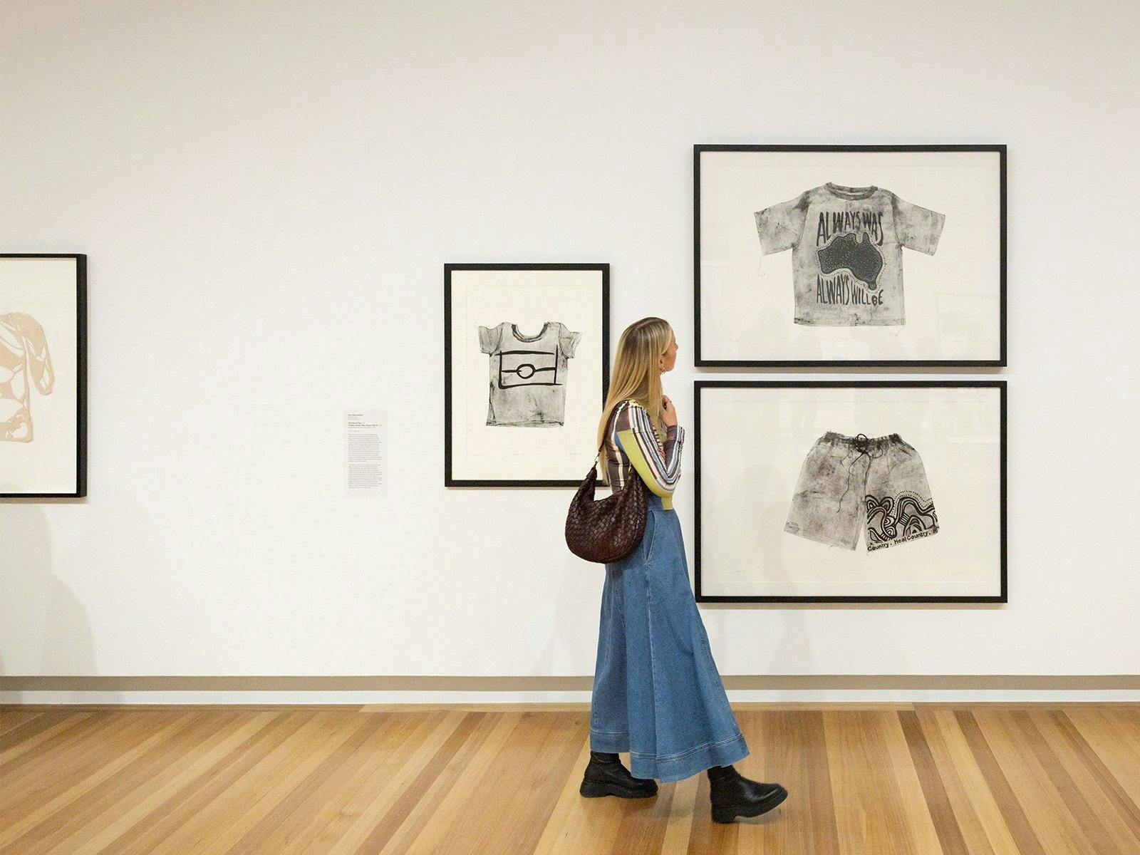 A person holding a brown handbag walks by a Gallery wall, looking at several artworks.