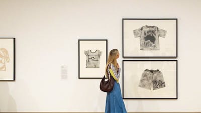 A person holding a brown handbag walks by a Gallery wall, looking at several artworks.