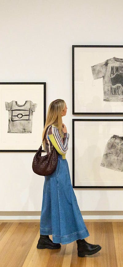 A person holding a brown handbag walks by a Gallery wall, looking at several artworks.