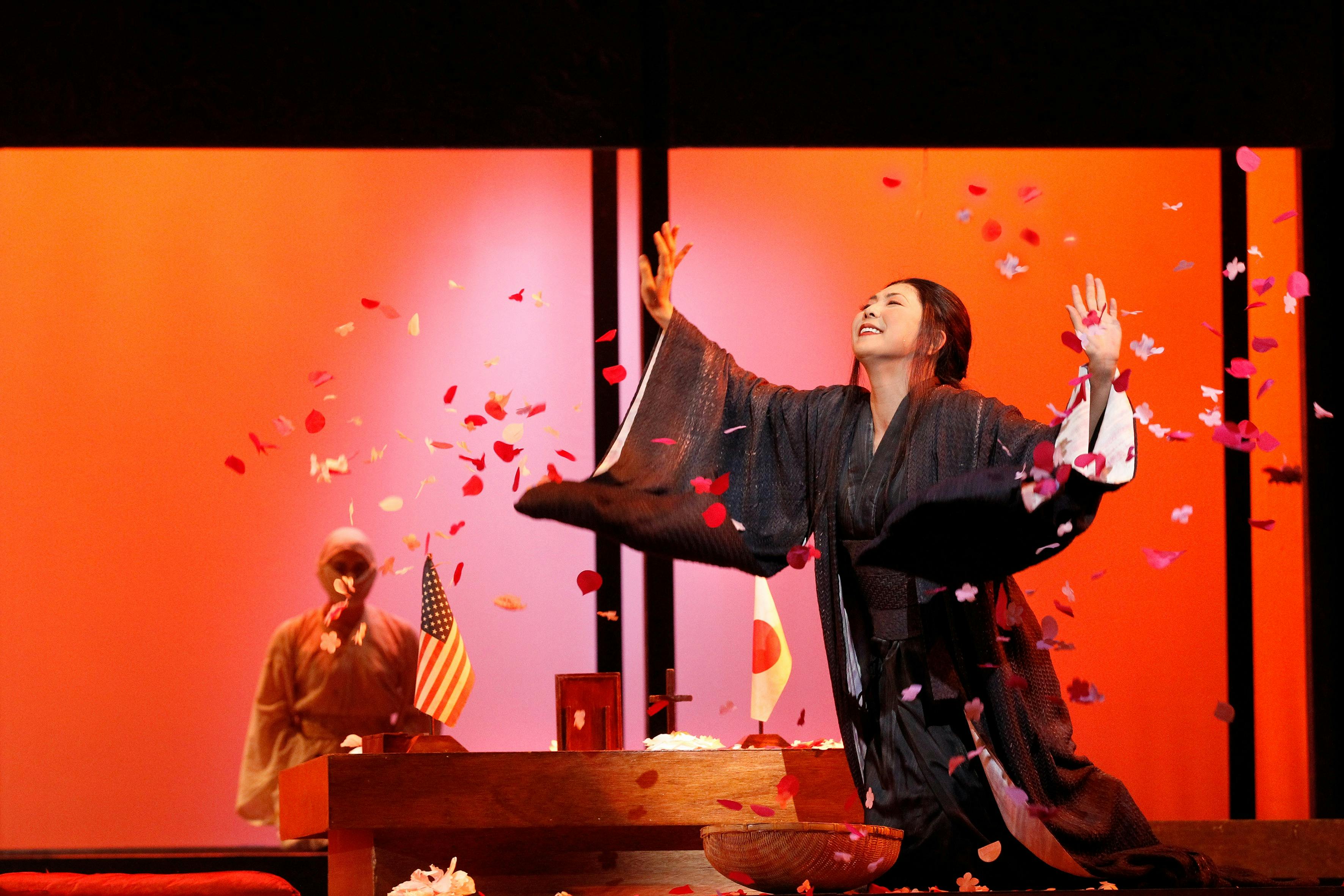 A woman in a dark kimono raises her arms as petals fall, with Japanese and US flags behind.