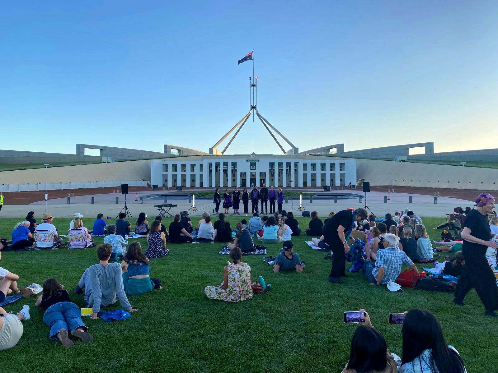 A group of people sitting on the lawns of Australian Parliament House watching a choir.