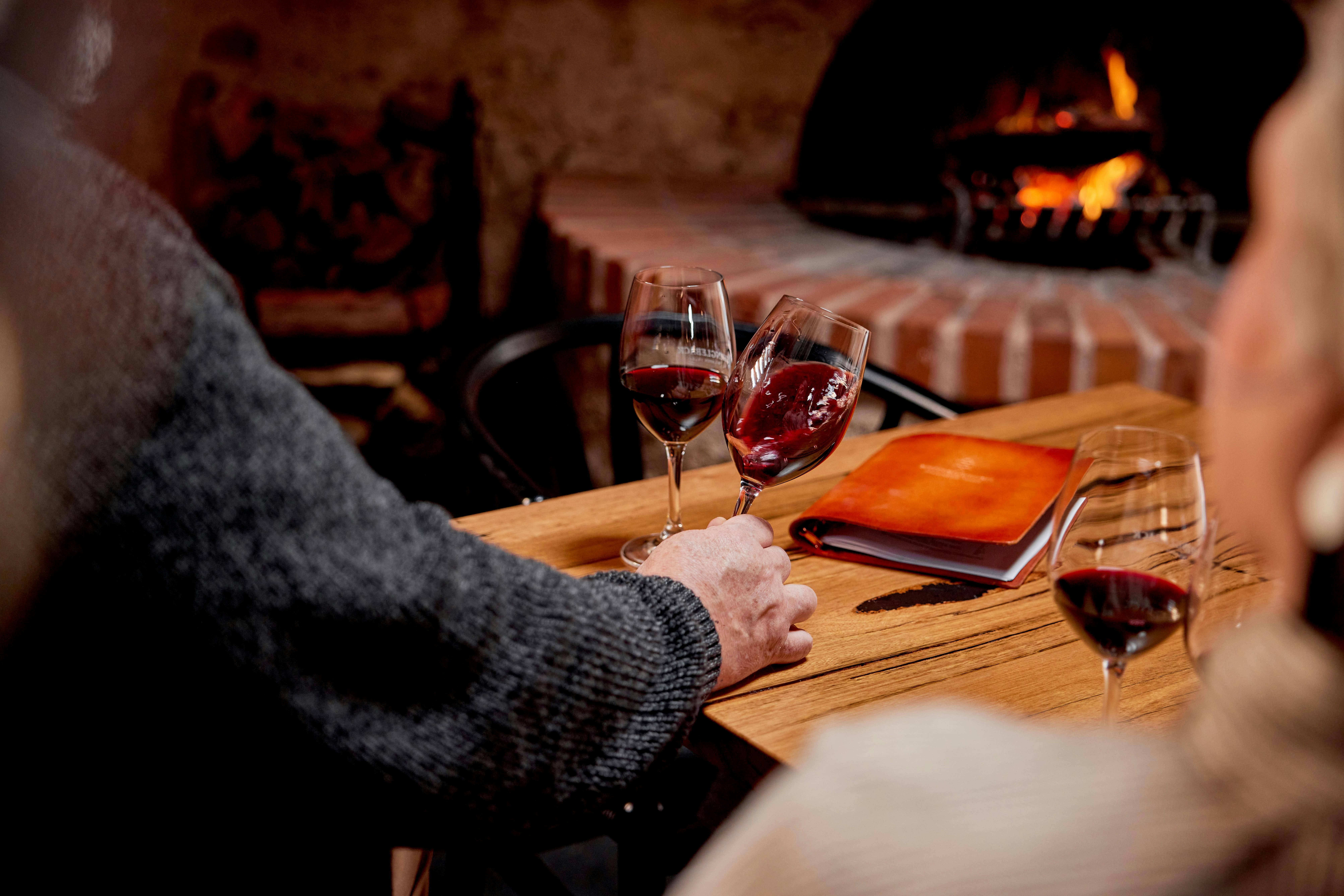 A man sits, with a glass of wine in hand, in front of the open fire in Shingleback's Cellar Door