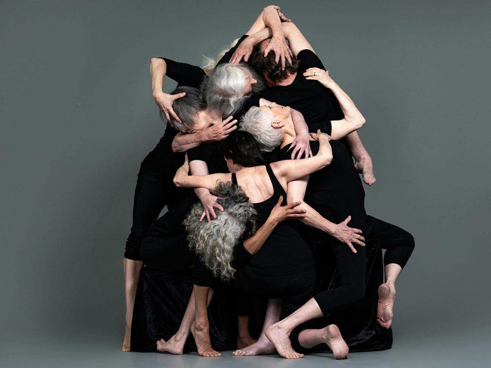 A group of dancers form a pyramid like abstract shape on a dark grey back ground.