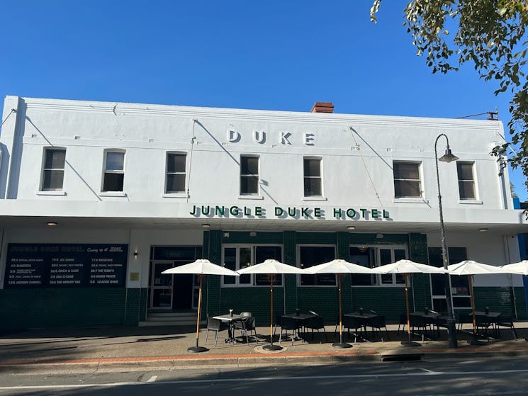 Image shows a white building called Jungle Duke Hotel, with tables and umbrellas outside