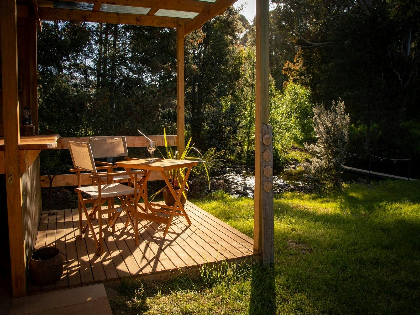 A deck area off the kitchen hut with  small table and 2 camp chairs. The deck overlooks the stream.
