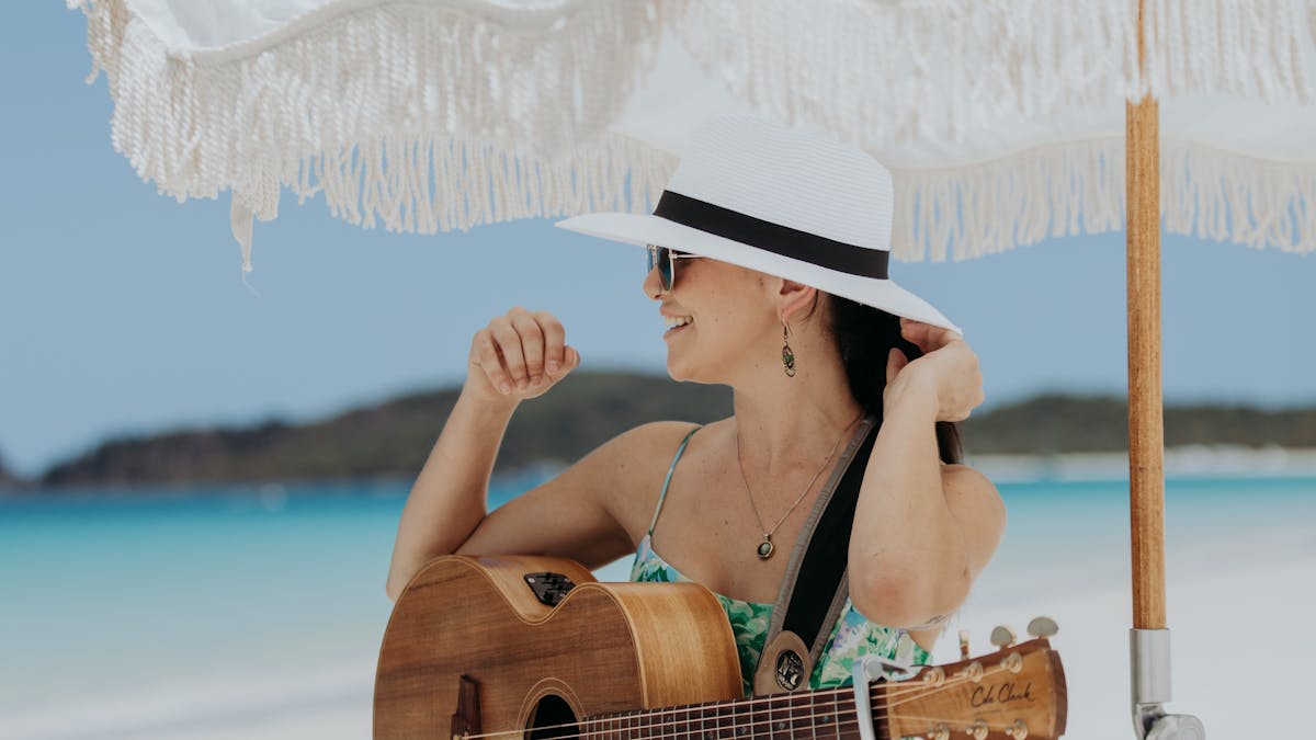 A female musician holding a guitar sitting on a stool on Whitehaven Beach underneath an  umbrella