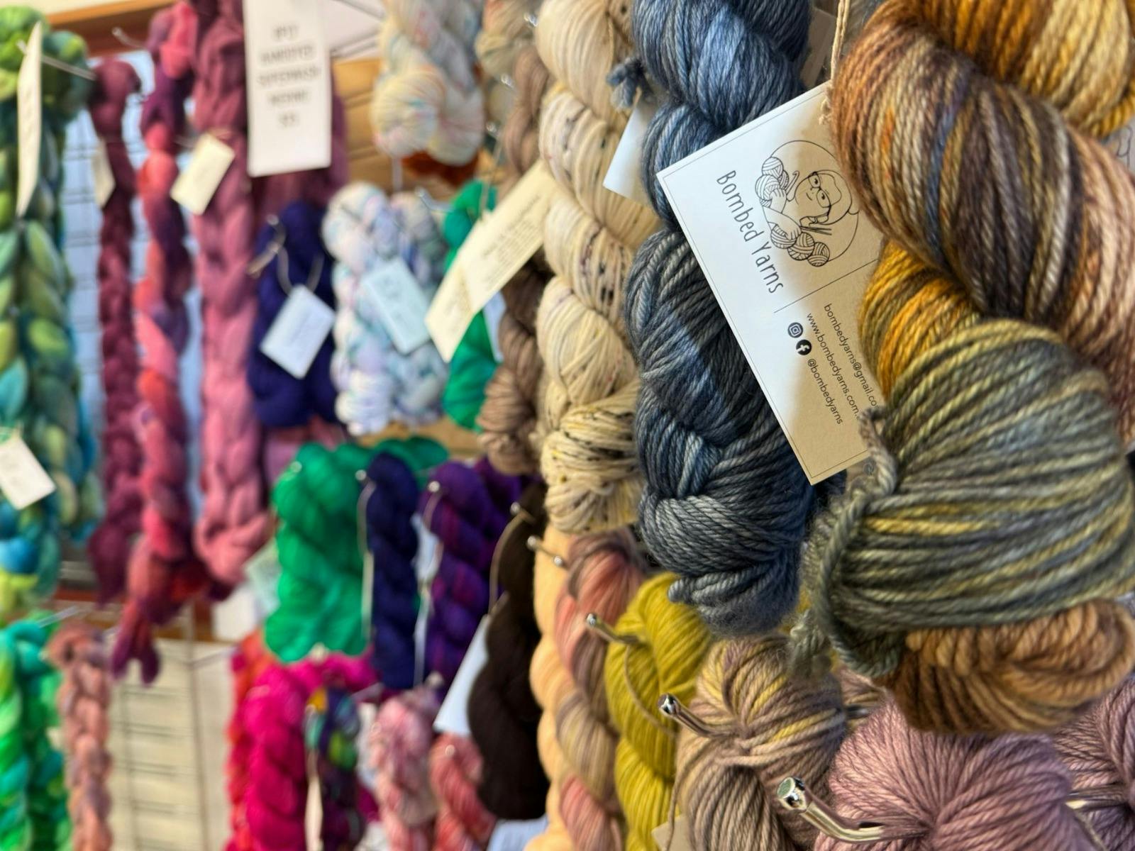 Skeins of colourful yarn hang from a rack at Bombed Yarns studio. There is a white label.