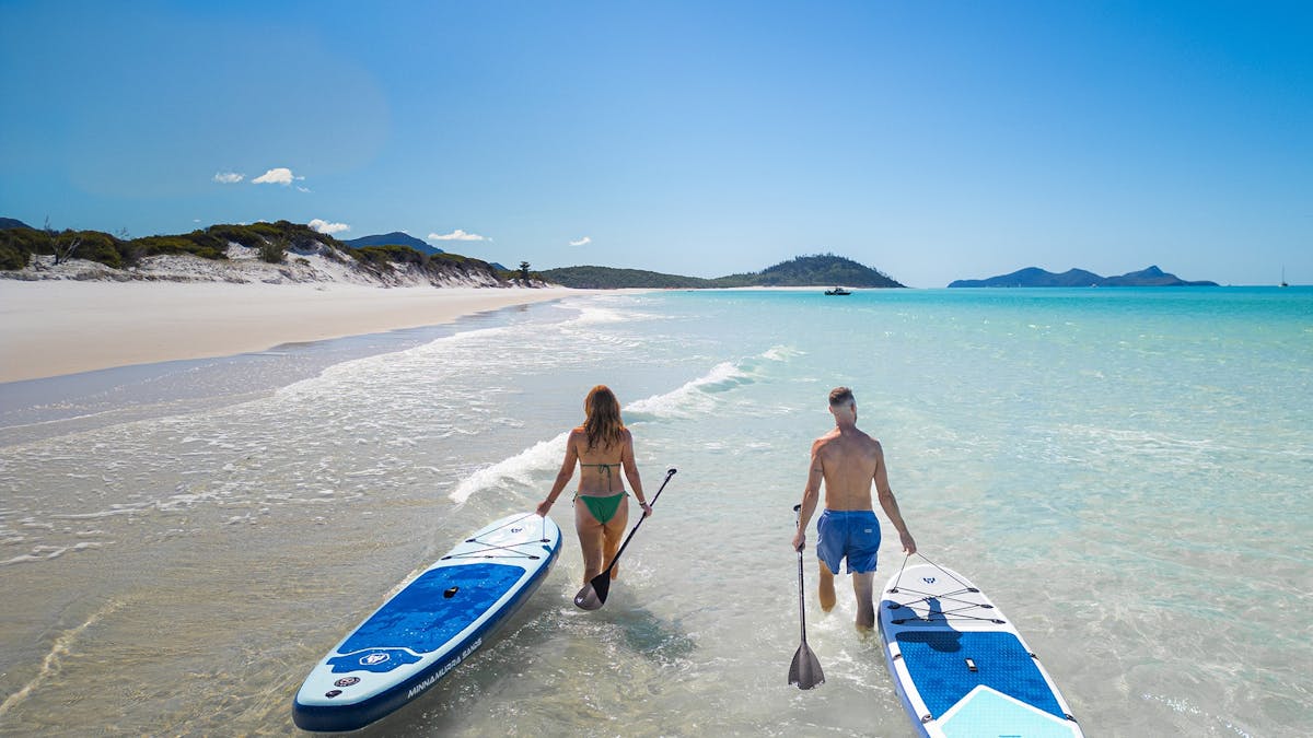 Two travellers carrying paddleboards through shallow turquoise water at Whitehaven Beach